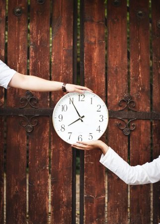 Two hands holding a clock in front of wooden gate.