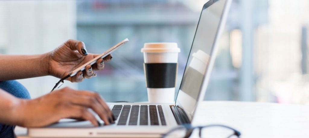 person typing into a laptop while holding a phone