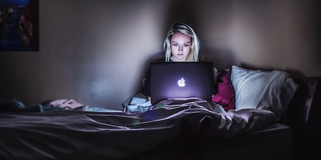 a woman sitting on the bed in the dark with a laptop