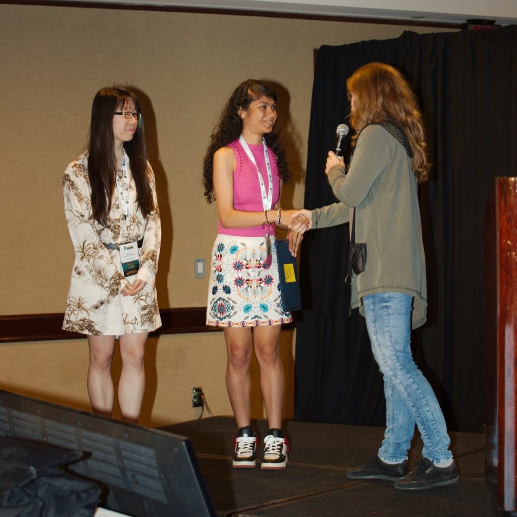 Photo of three people on stage, with the person in the middle, the award winner, receiving a handshake from the person on the right.