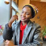 una mujer joven con una chaqueta de cuero gris sobre una camisa roja. Sonríe y mira por una ventana.