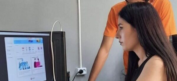 Teenage girl sitting at a table, using a desktop computer and a boy looking over her shoulder.
