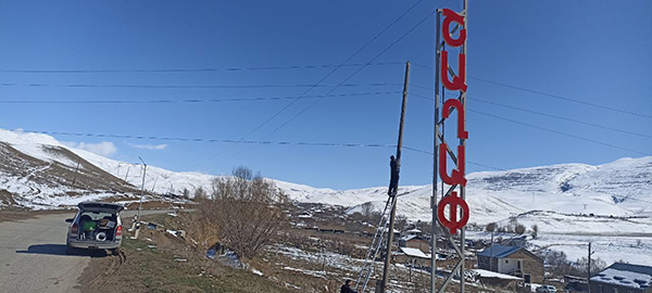 A la izquierda, una carretera con coche aparcado al lado y su maletero abierto. A la derecha, una gran torre con un letrero vertical en letra roja y una persona instalando una antena en la parte superior. Las montañas nevadas están en el fondo.