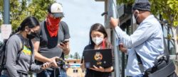 people wearing masks on a street, one holding an antenna, another one a laptop