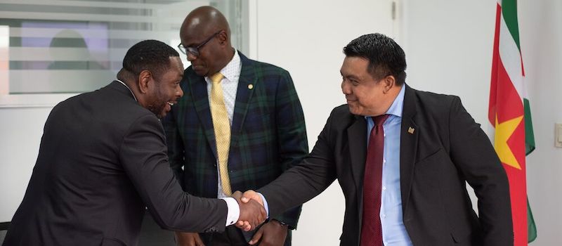 two men shaking hands smiling with Suriname flag in background