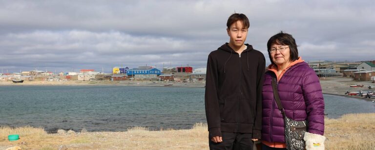 Two people standing in front of water and the hamlet of Ulukhaktok in the distance