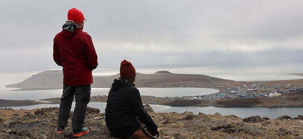 Two people on the top of a hill looking toward the horizon with the hamlet of Ulukhaktok and the ocean in the distance.