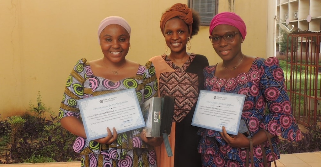 Three women proudly display their certificates in front of a building
