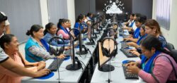 Women in blue vests sitting at computer monitors along two long tables.