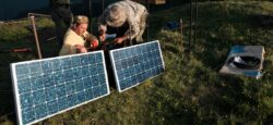Two men wiring a power inverter to solar panels amid green mountains.