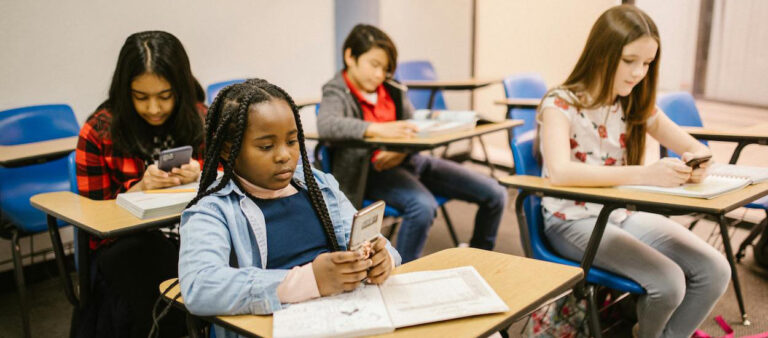 Four children sit at school desks and use smartphones