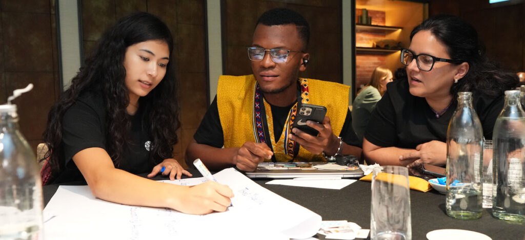 Three people sit at a table and look at a piece of paper, which one person is writing on.