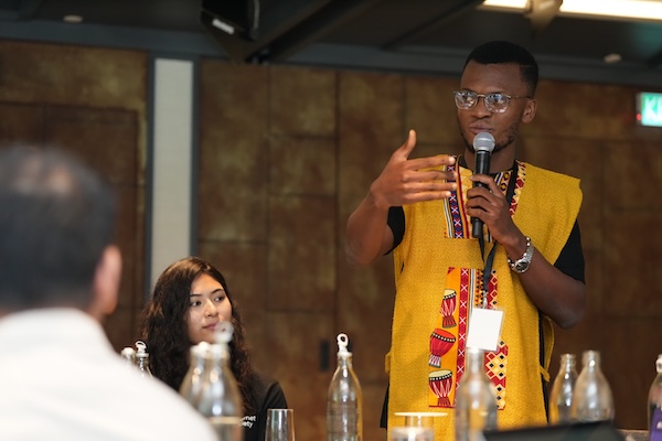 A man in a yellow shirt stands at a podium, delivering a speech
