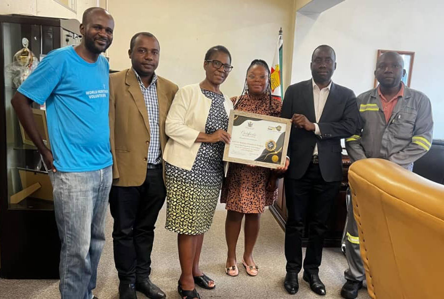 Six people stand in a row, the two in the center holding a certificate