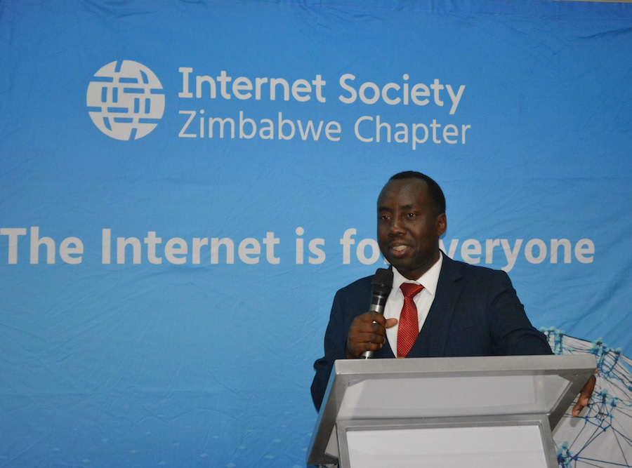 A man in a suit stands at a podium and speaks. Behind him, a blue sign shows the Internet Society Zimbabwe Chapter logo and the phrase 'the Internet is for everyone.'