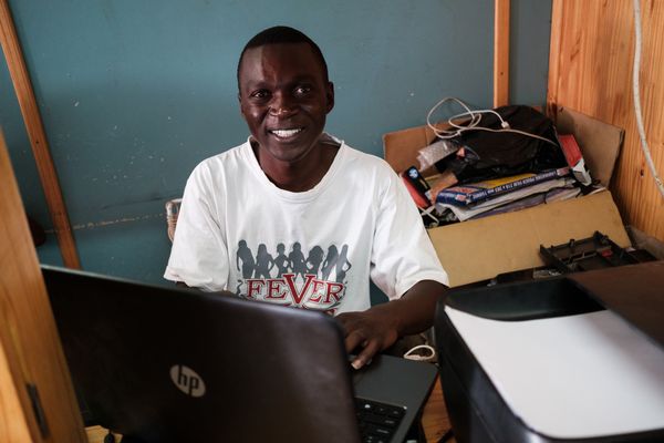 A man smiles while seated at a desk, working on a laptop