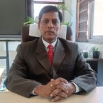 a man in a suit with a red tie sitting at his desk posing for a photo