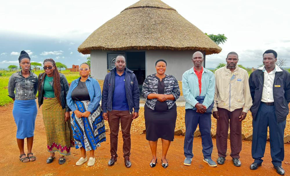 Eight people stand together side-by-side in front of a small building in rural Zimbabwe.