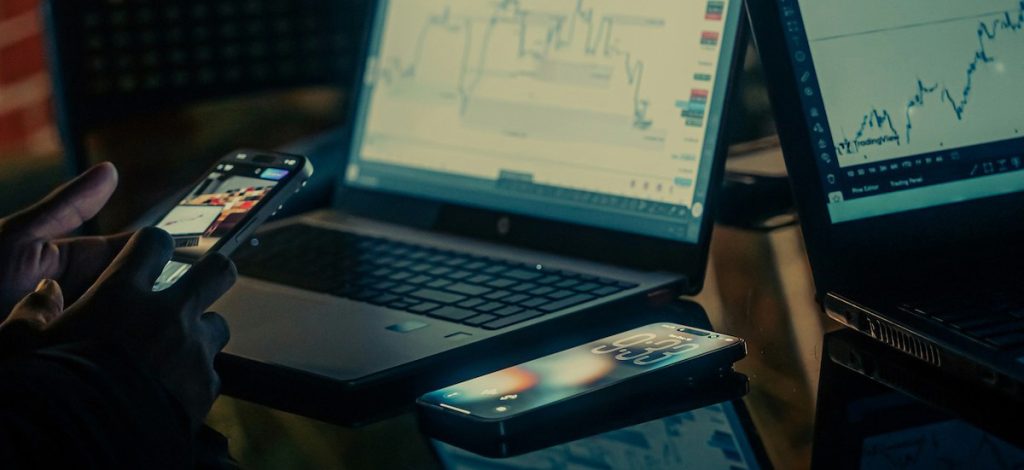 Two computers and a phone sit on a table, hands use another phone.