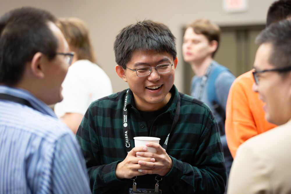 A man stands, smiles, and holds a cup of coffee while conversing at the NDSS Symposium 2026