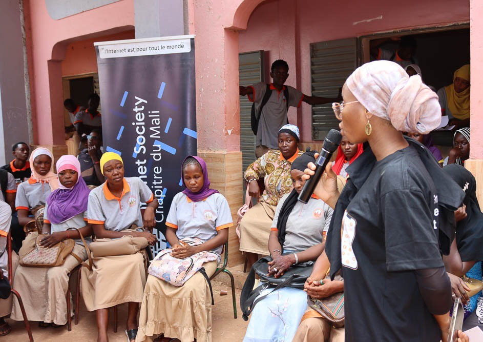 A woman stands and speaks into a microphone, talking to a seated group of women.