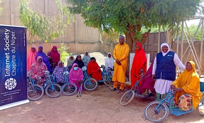 Several participants are seated on blue tricycles, while others stand nearby.