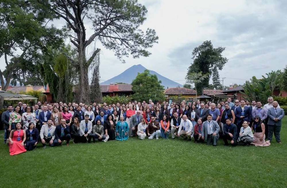 A large group of people kneel and stand together outside, with trees and a mountain in the background.