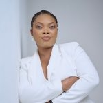 A woman in a white suit leans against a wall