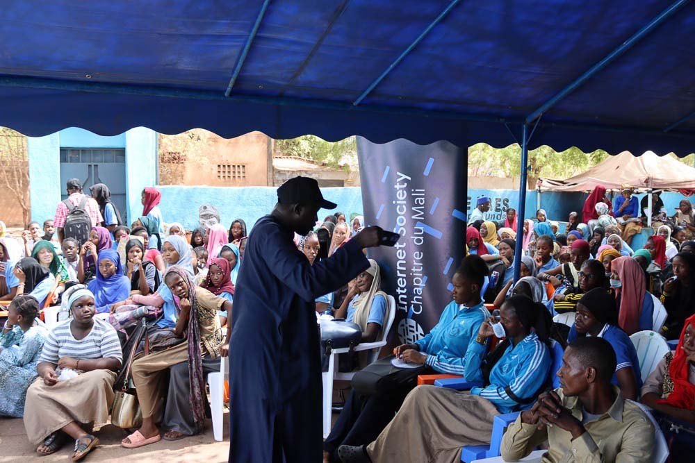 A man stands and speaks to a large seated group outside next to an Internet Society Mali Chapter sign.