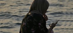 A woman looks down at her phone with water in the background