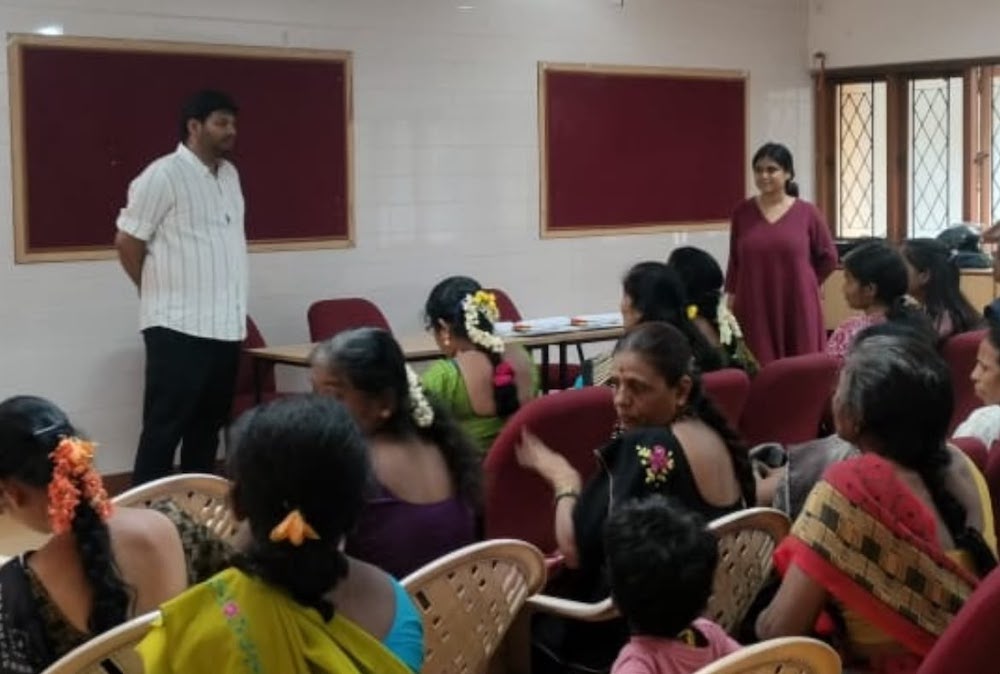 A man in a white shirt stands in a room in front of a seated group.