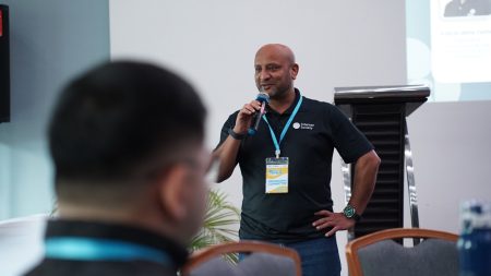 A man wearing a black shirt presents information to an audience with a microphone
