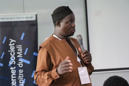 A man stands and speaks into a microphone next to an Internet Society Mali Chapter sign.