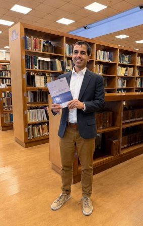 A man stands in a library, holding a book.