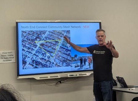 A man stands and speaks, pointing at a powerpoint.