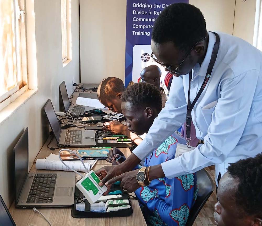 A man in a light blue button-up shirt stands and handles electronic equipment, pointing things out to young, seated trainees.