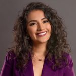 A woman with curly hair wearing a purple blazer, smiling.