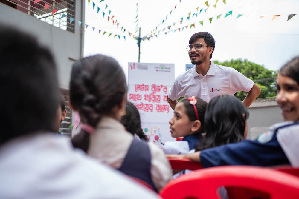 A man in a white shirt stands outside in front of a seated group.